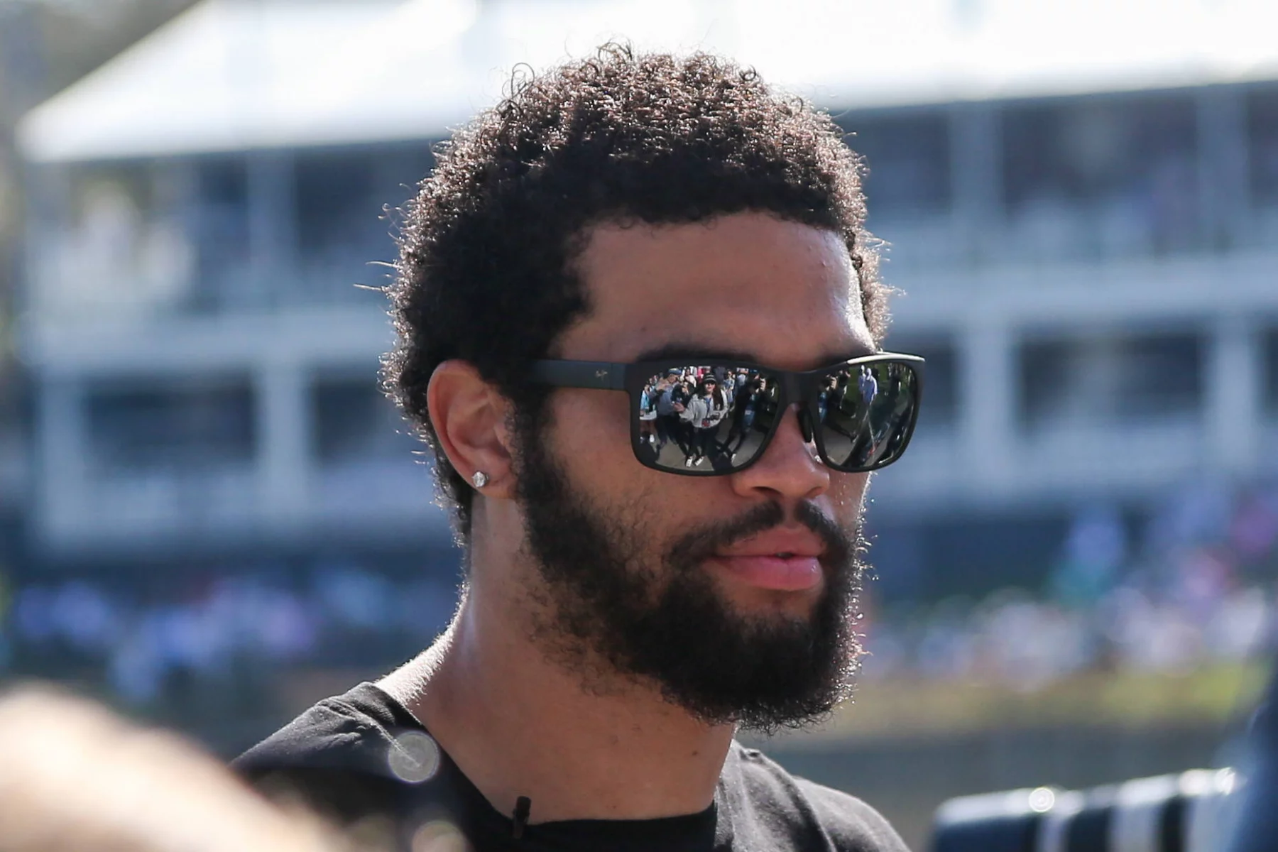 FootballR - NFL - Start, Sit und Drop - Ein Mann mit gepflegtem Bart und Sonnenbrille steht draußen, Sonnenlicht beleuchtet sein lockiges Haar, während während des Pro Day von Caleb Williams die Umgebung in seiner Sonnenbrille glitzert. Diese Beschreibung wurde automatisch generiert.