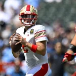 FootballR - NFL - Matt Corral von den Stallions, ein konzentrierter Quarterback in weiß-roter Ausrüstung, sucht während eines intensiven UFL-Auftakt-Footballspiels, bei dem die Verteidiger dicht auf den Ball kommen, nach einem offenen Teamkollegen, der den Ball weiterspielen kann Diese Beschreibung wurde automatisch generiert.