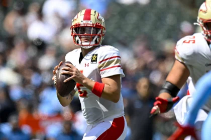 FootballR - NFL - Matt Corral von den Stallions, ein konzentrierter Quarterback in weiß-roter Ausrüstung, sucht während eines intensiven UFL-Auftakt-Footballspiels, bei dem die Verteidiger dicht auf den Ball kommen, nach einem offenen Teamkollegen, der den Ball weiterspielen kann Diese Beschreibung wurde automatisch generiert.