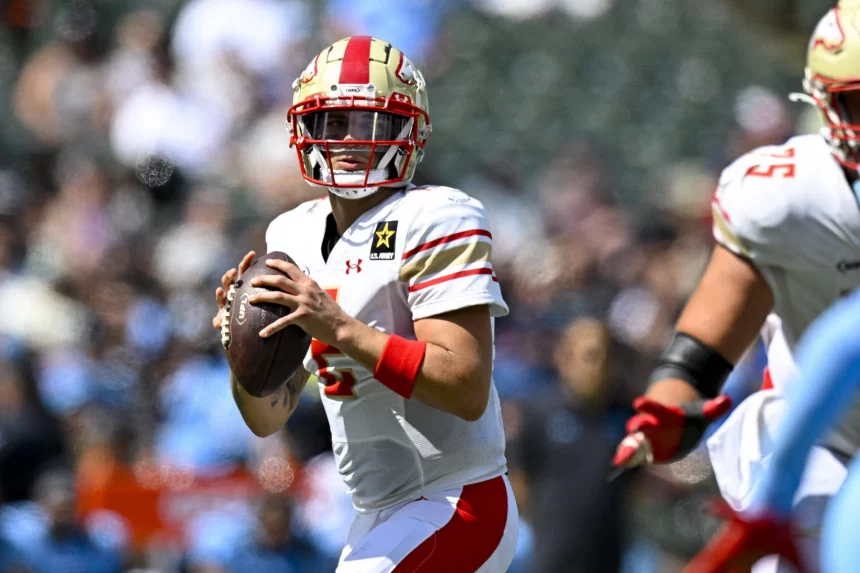 FootballR - NFL - Matt Corral von den Stallions, ein konzentrierter Quarterback in weiß-roter Ausrüstung, sucht während eines intensiven UFL-Auftakt-Footballspiels, bei dem die Verteidiger dicht auf den Ball kommen, nach einem offenen Teamkollegen, der den Ball weiterspielen kann Diese Beschreibung wurde automatisch generiert.