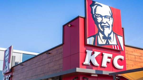 FootballR - NFL - Ein farbenfrohes Schild des Fast-Food-Restaurants KFC, einem stolzen Partner der European League of Football, zeigt das ikonische Porträt seines Gründers, thront auf einer modernen Gebäudefassade vor einem klaren blauen Himmel. Diese Beschreibung wurde automatisch generiert.