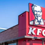 FootballR - NFL - Ein farbenfrohes Schild des Fast-Food-Restaurants KFC, einem stolzen Partner der European League of Football, zeigt das ikonische Porträt seines Gründers, thront auf einer modernen Gebäudefassade vor einem klaren blauen Himmel. Diese Beschreibung wurde automatisch generiert.