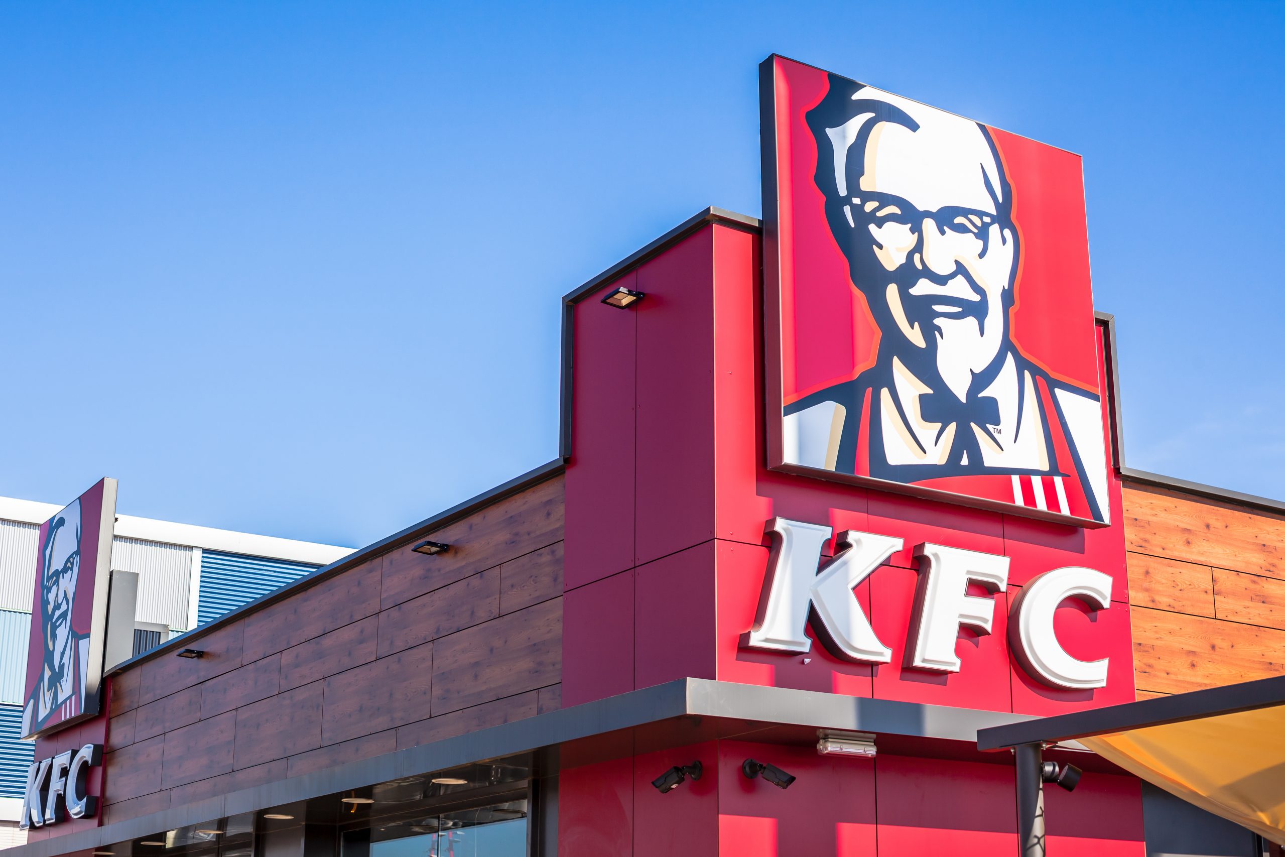 FootballR - NFL - Ein farbenfrohes Schild des Fast-Food-Restaurants KFC, einem stolzen Partner der European League of Football, zeigt das ikonische Porträt seines Gründers, thront auf einer modernen Gebäudefassade vor einem klaren blauen Himmel. Diese Beschreibung wurde automatisch generiert.
