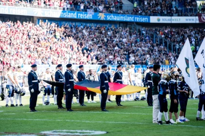 FootballR - NFL - An einem sonnigen Tag tragen uniformierte Bundeswehrangehörige vor einem vollen Stadionpublikum eine große deutsche Flagge über ein Feld, was an eine formelle Zeremonie oder ein Eröffnungsritual eines Spiels der European League of Football erinnert. Diese Beschreibung wurde automatisch generiert.