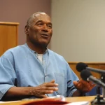 FootballR - NFL - Ein Mann in blauer Gefängnisuniform, identifiziert als die umstrittene Figur und Football-Legende O.J. Simpson, sitzt an einem Tisch mit Mikrofonen und lächelt während einer öffentlichen Rede in einem Raum mit Holz Diese Beschreibung wurde automatisch generiert. O.J. Simpson