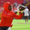 FootballR - NFL - Rashee Rice, ein konzentrierter Footballspieler, trägt einen roten Kapuzenpullover und Handschuhe und übt auf dem Feld das Fangen eines Passes. Bei einer Aufwärmübung vor einem Spiel zeigt er Konzentration und sportliches Können. Diese Beschreibung wurde automatisch generiert.