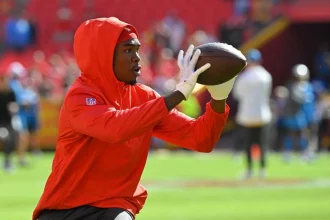 FootballR - NFL - Rashee Rice, ein konzentrierter Footballspieler, trägt einen roten Kapuzenpullover und Handschuhe und übt auf dem Feld das Fangen eines Passes. Bei einer Aufwärmübung vor einem Spiel zeigt er Konzentration und sportliches Können. Diese Beschreibung wurde automatisch generiert.