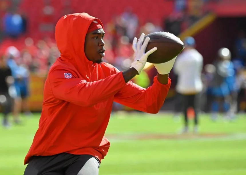 FootballR - NFL - Rashee Rice, ein konzentrierter Footballspieler, trägt einen roten Kapuzenpullover und Handschuhe und übt auf dem Feld das Fangen eines Passes. Bei einer Aufwärmübung vor einem Spiel zeigt er Konzentration und sportliches Können. Diese Beschreibung wurde automatisch generiert.