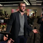 FootballR - NFL Andrew Luck - Ein Mann mit Brille und grauem Blazer geht mit offenem Lächeln durch eine Menschenmenge bei einer offiziellen Veranstaltung in Indianapolis und gestikuliert mit offenen Händen in Richtung Reporter, die Mikrofone halten, in einem Raum mit Abendessen Diese Beschreibung wurde automatisch generiert.