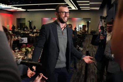 FootballR - NFL Andrew Luck - Ein Mann mit Brille und grauem Blazer geht mit offenem Lächeln durch eine Menschenmenge bei einer offiziellen Veranstaltung in Indianapolis und gestikuliert mit offenen Händen in Richtung Reporter, die Mikrofone halten, in einem Raum mit Abendessen Diese Beschreibung wurde automatisch generiert.