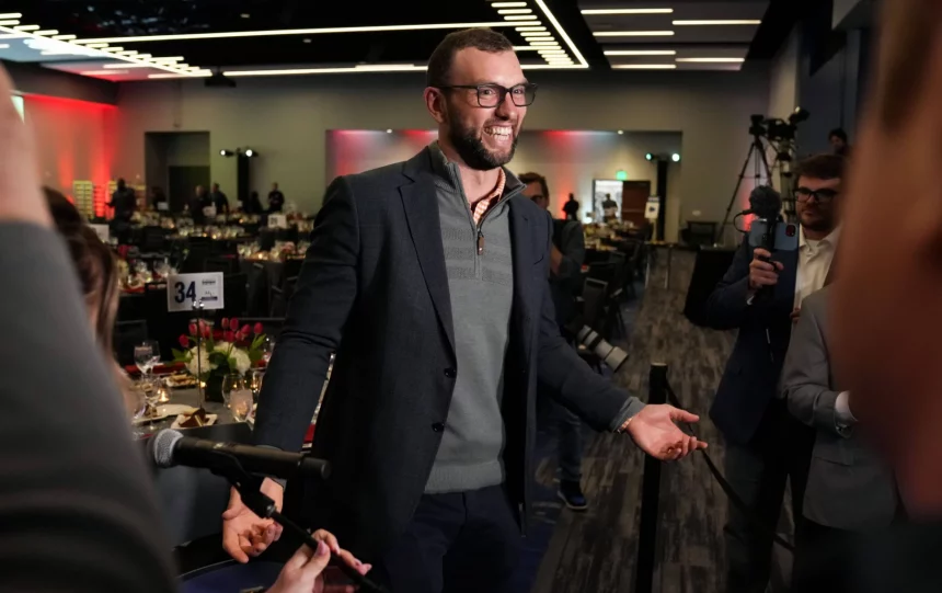 FootballR - NFL Andrew Luck - Ein Mann mit Brille und grauem Blazer geht mit offenem Lächeln durch eine Menschenmenge bei einer offiziellen Veranstaltung in Indianapolis und gestikuliert mit offenen Händen in Richtung Reporter, die Mikrofone halten, in einem Raum mit Abendessen Diese Beschreibung wurde automatisch generiert.