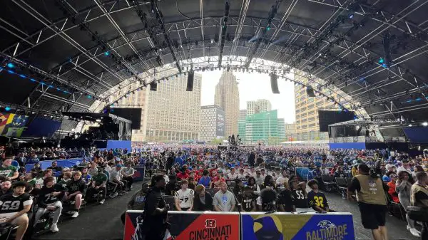 FootballR - NFL - Detroit - Eine große Menschenmenge versammelte sich in einem weitläufigen Veranstaltungsort mit hoher, gewölbter Decke für den NFL Draft 2024. Auf der Bühne hängen Banner der Cincinnati Bengals und Baltimore Ravens. Eine Stadtkulisse fügt Diese Beschreibung wurde automatisch generiert.