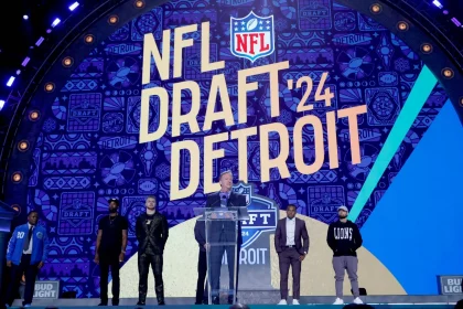 FootballR - NFL - Ein NFL Draft 2024 Event in Detroit mit einer großen, lebendigen Bühne, auf der in fetten Buchstaben „NFL Draft '24 Detroit“ steht. Ein Redner steht an einem Podium, flankiert von Figuren in Detroit Diese Beschreibung wurde automatisch generiert.