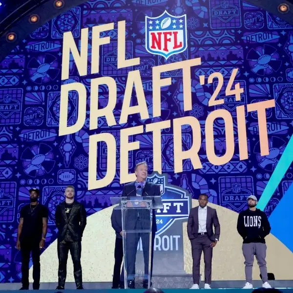 FootballR - NFL - Ein NFL Draft 2024 Event in Detroit mit einer großen, lebendigen Bühne, auf der in fetten Buchstaben „NFL Draft '24 Detroit“ steht. Ein Redner steht an einem Podium, flankiert von Figuren in Detroit Diese Beschreibung wurde automatisch generiert.