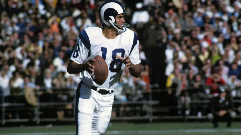 FootballR - NFL - Ein Quarterback in weiß-blauer Uniform mit der Nummer 18, identifiziert als NFL MVP Roman Gabriel, sucht während eines sonnigen Footballspiels nach einem freien Receiver. Er hält den Football in seiner rechten Diese Beschreibung wurde automatisch generiert.