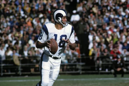 FootballR - NFL - Ein Quarterback in weiß-blauer Uniform mit der Nummer 18, identifiziert als NFL MVP Roman Gabriel, sucht während eines sonnigen Footballspiels nach einem freien Receiver. Er hält den Football in seiner rechten Diese Beschreibung wurde automatisch generiert.