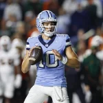 FootballR - NFL - Drake Maye - Ein konzentrierter Quarterback der New England Patriots in der blau-weißen Uniform mit der Nummer 10 sucht während eines nächtlichen Footballspiels das Spielfeld nach einem freien Receiver ab. Im Hintergrund sind die Spieler der gegnerischen Mannschaft zu sehen. Diese Beschreibung wurde automatisch generiert.