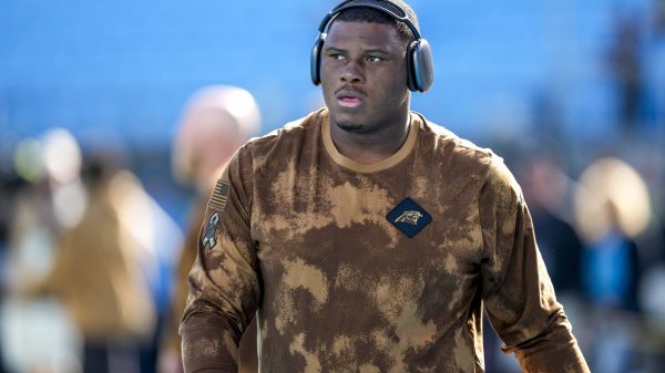 FootballR - NFL - Derrick Brown - Ein konzentrierter Athlet in einem Camouflage-Kapuzenpullover der Carolina Panthers und mit Kopfhörern schreitet über das Feld und strahlt Entschlossenheit und Bereitschaft für das bevorstehende Spiel aus. Diese Beschreibung wurde automatisch generiert.