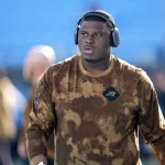FootballR - NFL - Derrick Brown - Ein konzentrierter Athlet in einem Camouflage-Kapuzenpullover der Carolina Panthers und mit Kopfhörern schreitet über das Feld und strahlt Entschlossenheit und Bereitschaft für das bevorstehende Spiel aus. Diese Beschreibung wurde automatisch generiert.