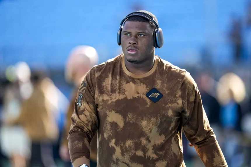 FootballR - NFL - Derrick Brown - Ein konzentrierter Athlet in einem Camouflage-Kapuzenpullover der Carolina Panthers und mit Kopfhörern schreitet über das Feld und strahlt Entschlossenheit und Bereitschaft für das bevorstehende Spiel aus. Diese Beschreibung wurde automatisch generiert.