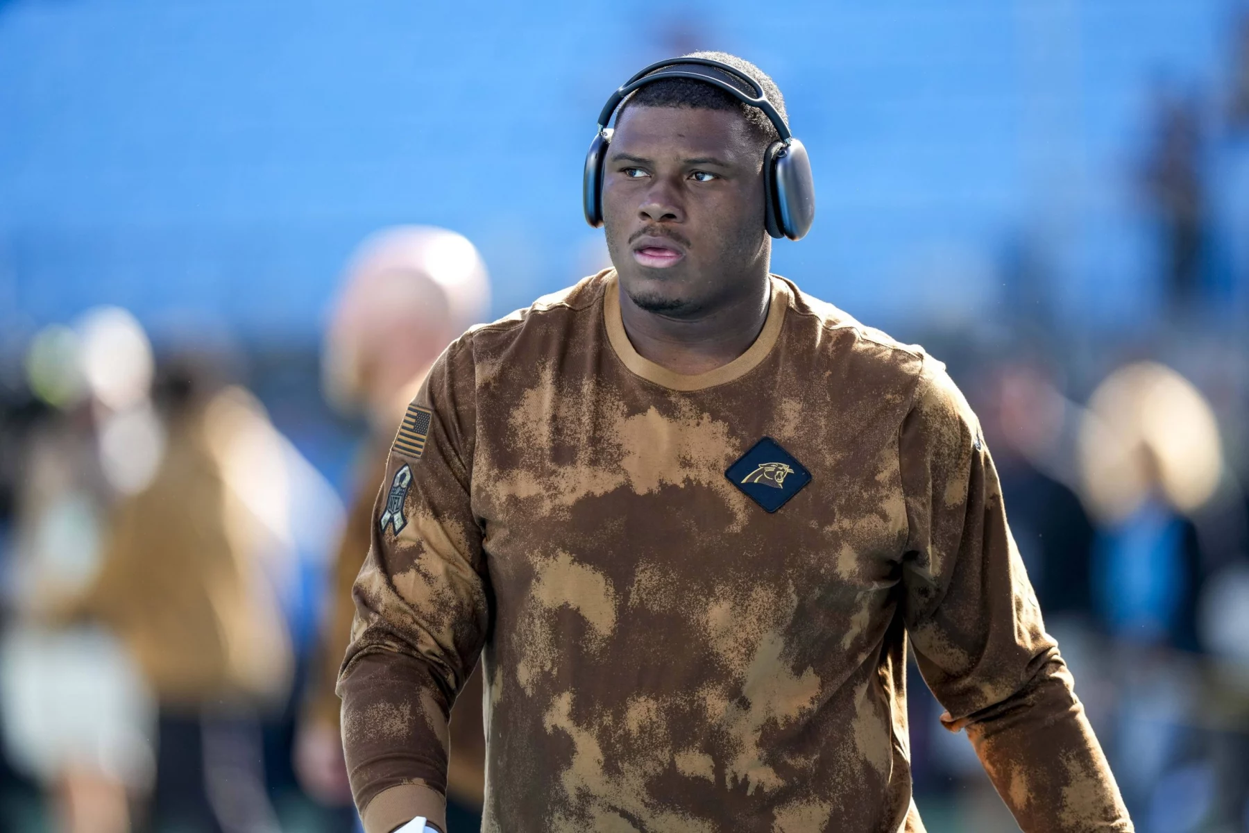 FootballR - NFL - Derrick Brown - Ein konzentrierter Athlet in einem Camouflage-Kapuzenpullover der Carolina Panthers und mit Kopfhörern schreitet über das Feld und strahlt Entschlossenheit und Bereitschaft für das bevorstehende Spiel aus. Diese Beschreibung wurde automatisch generiert.