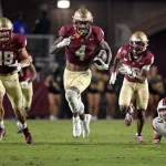 FootballR - NFL - Drei Footballspieler des Staates Florida in kastanienbraunen und goldenen Uniformen laufen während eines Nachtspiels, während hinter ihnen ein Gegner in Weiß und Orange auf dem Boden liegt, darunter WR Keon Coleman. Diese Beschreibung wurde automatisch generiert.