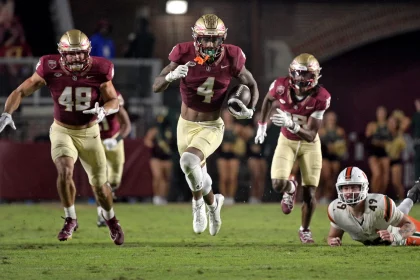 FootballR - NFL - Drei Footballspieler des Staates Florida in kastanienbraunen und goldenen Uniformen laufen während eines Nachtspiels, während hinter ihnen ein Gegner in Weiß und Orange auf dem Boden liegt, darunter WR Keon Coleman. Diese Beschreibung wurde automatisch generiert.