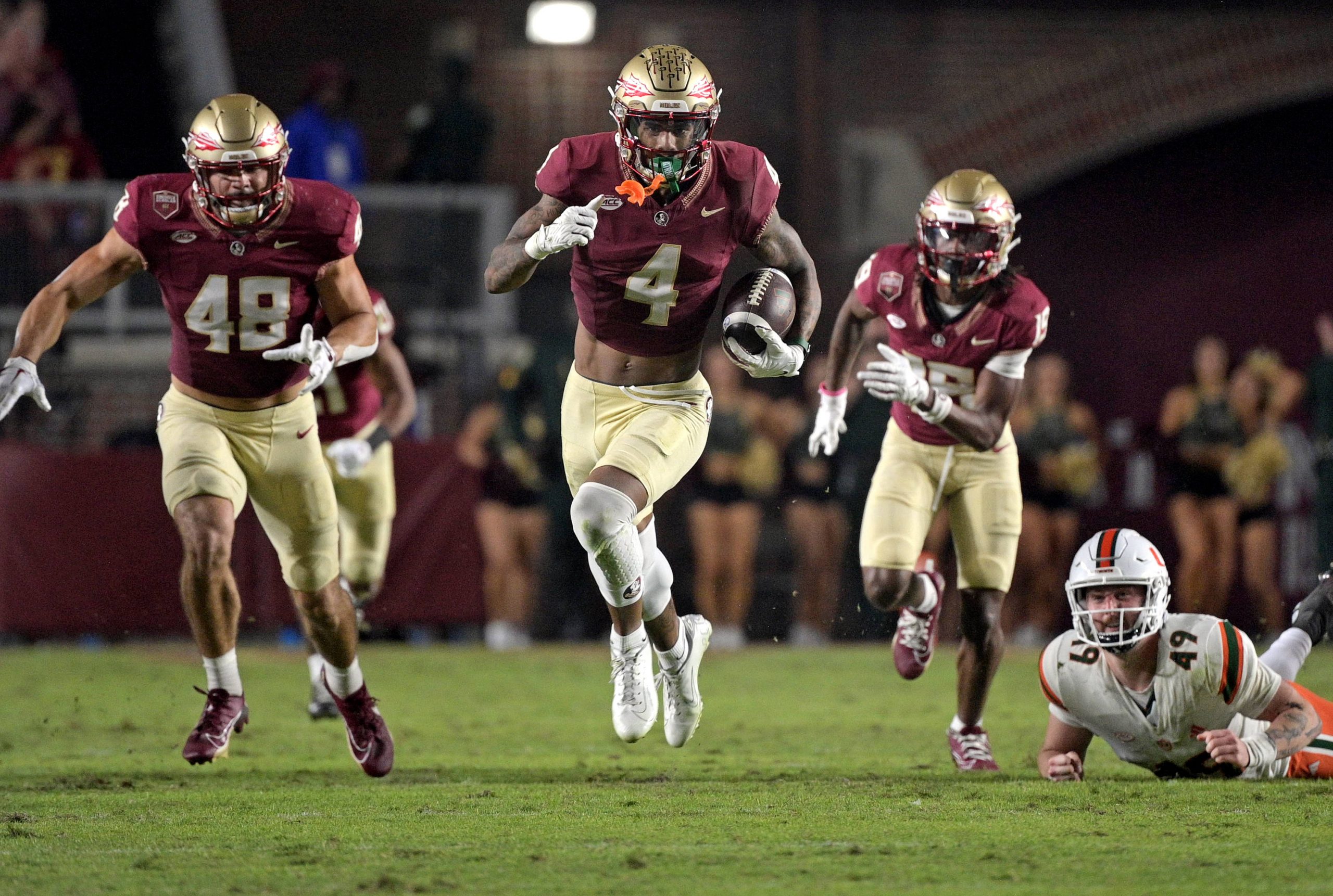 FootballR - NFL - Drei Footballspieler des Staates Florida in kastanienbraunen und goldenen Uniformen laufen während eines Nachtspiels, während hinter ihnen ein Gegner in Weiß und Orange auf dem Boden liegt, darunter WR Keon Coleman. Diese Beschreibung wurde automatisch generiert.