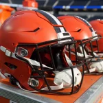 FootballR - NFL - Eine Reihe glänzender, roter American-Football-Helme mit schützenden Gesichtsmasken, die für die Helmsicherheit entwickelt wurden, erwarten die Spieler am Spielfeldrand und signalisieren die Vorbereitungen für ein bevorstehendes Spiel. Diese Beschreibung wurde automatisch generiert.