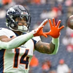FootballR - NFL - Courtland Sutton, ein Spieler der Denver Broncos mit der Nummer 14, streckt während eines Spiels die Hand aus, um einen Football zu fangen. Er ist hochkonzentriert und seine orangefarbenen Handschuhe stechen von seiner blau-weißen Uniform hervor. Diese Beschreibung wurde automatisch generiert.