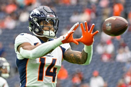 FootballR - NFL - Courtland Sutton, ein Spieler der Denver Broncos mit der Nummer 14, streckt während eines Spiels die Hand aus, um einen Football zu fangen. Er ist hochkonzentriert und seine orangefarbenen Handschuhe stechen von seiner blau-weißen Uniform hervor. Diese Beschreibung wurde automatisch generiert.