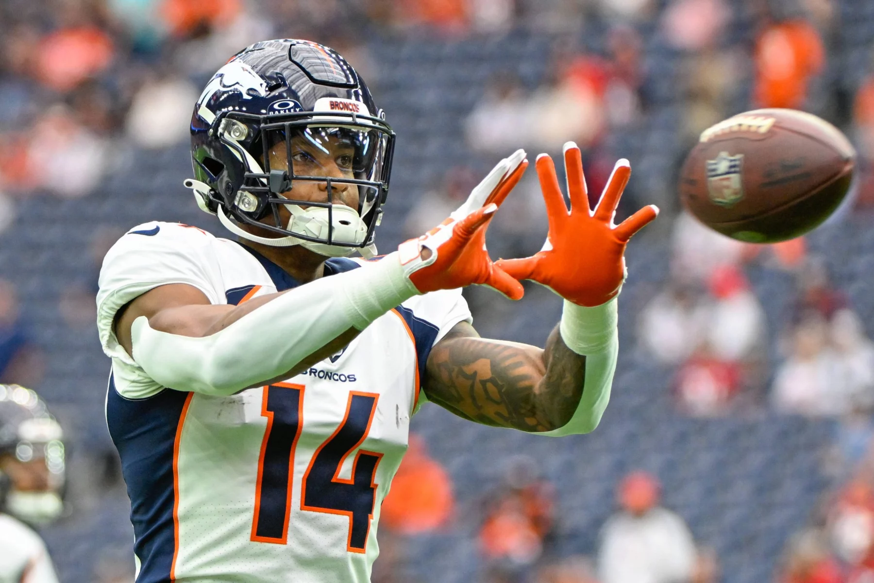 FootballR - NFL - Courtland Sutton, ein Spieler der Denver Broncos mit der Nummer 14, streckt während eines Spiels die Hand aus, um einen Football zu fangen. Er ist hochkonzentriert und seine orangefarbenen Handschuhe stechen von seiner blau-weißen Uniform hervor. Diese Beschreibung wurde automatisch generiert.
