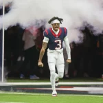 FootballR - NFL - Tank Dell - Ein Footballspieler in der weißen Uniform mit der Nummer 3 und roten und blauen Details rennt aufgeregt durch eine weiße Rauchwolke auf das Spielfeld, in einem voll besetzten Stadion. Sein Haar ist in auffälligen Diese Beschreibung wurde automatisch generiert.