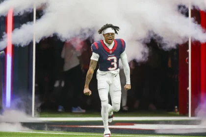 FootballR - NFL - Tank Dell - Ein Footballspieler in der weißen Uniform mit der Nummer 3 und roten und blauen Details rennt aufgeregt durch eine weiße Rauchwolke auf das Spielfeld, in einem voll besetzten Stadion. Sein Haar ist in auffälligen Diese Beschreibung wurde automatisch generiert.