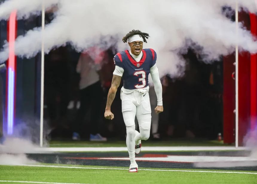 FootballR - NFL - Tank Dell - Ein Footballspieler in der weißen Uniform mit der Nummer 3 und roten und blauen Details rennt aufgeregt durch eine weiße Rauchwolke auf das Spielfeld, in einem voll besetzten Stadion. Sein Haar ist in auffälligen Diese Beschreibung wurde automatisch generiert.