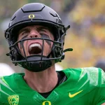 FootballR - NFL - Ein Footballspieler, Bo Nix, in einer leuchtend grün-blauen Seahawks-Uniform mit einer auffälligen Nummer 10, blickt mit angespanntem Gesichtsausdruck nach oben, sein Mundschutz ist sichtbar, vor einem verschwommenen Hintergrund mit einer Menschenmenge Diese Beschreibung wurde automatisch generiert.