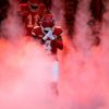 FootballR - NFL Rashee Rice - Ein Wide Receiver der Kansas City Chiefs in einer roten Uniform mit der Nummer 14 taucht dramatisch aus einer roten Rauchwolke auf, im Hintergrund sind teilweise Teamkollegen zu sehen, was einen intensiven Moment vor dem Spiel auf dem Diese Beschreibung wurde automatisch generiert.