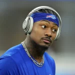 FootballR - NFL - Stefon Diggs - Ein konzentrierter Sportler in einem blauen T-Shirt der Buffalo Bills hört Musik über seine Over-Ear-Kopfhörer, während er eine goldene Kette trägt und sich auf ein Spiel vorbereitet. Damit zeigt er seine Loyalität, bevor er über Free zu den Texans wechselt. Diese Beschreibung wurde automatisch generiert.