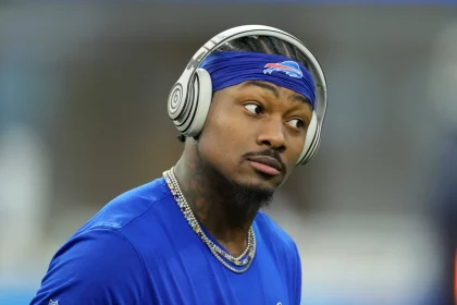 FootballR - NFL - Stefon Diggs - Ein konzentrierter Sportler in einem blauen T-Shirt der Buffalo Bills hört Musik über seine Over-Ear-Kopfhörer, während er eine goldene Kette trägt und sich auf ein Spiel vorbereitet. Damit zeigt er seine Loyalität, bevor er über Free zu den Texans wechselt. Diese Beschreibung wurde automatisch generiert.