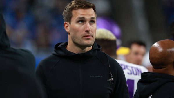 FootballR - NFL Kirk Cousins - Ein konzentrierter Quarterback in einem schwarzen Kapuzenpullover mit einem „Vikings“-Logo steht am Spielfeldrand eines Sportstadions, umgeben von anderen Menschen, hinter ihm eine Menschenmenge auf den Tribünen. Diese Beschreibung wurde automatisch generiert.