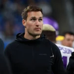 FootballR - NFL Kirk Cousins - Ein konzentrierter Quarterback in einem schwarzen Kapuzenpullover mit einem „Vikings“-Logo steht am Spielfeldrand eines Sportstadions, umgeben von anderen Menschen, hinter ihm eine Menschenmenge auf den Tribünen. Diese Beschreibung wurde automatisch generiert.