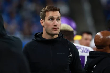 FootballR - NFL Kirk Cousins - Ein konzentrierter Quarterback in einem schwarzen Kapuzenpullover mit einem „Vikings“-Logo steht am Spielfeldrand eines Sportstadions, umgeben von anderen Menschen, hinter ihm eine Menschenmenge auf den Tribünen. Diese Beschreibung wurde automatisch generiert.