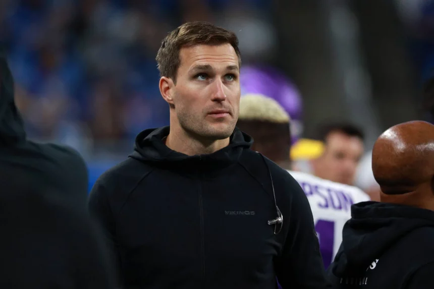 FootballR - NFL Kirk Cousins - Ein konzentrierter Quarterback in einem schwarzen Kapuzenpullover mit einem „Vikings“-Logo steht am Spielfeldrand eines Sportstadions, umgeben von anderen Menschen, hinter ihm eine Menschenmenge auf den Tribünen. Diese Beschreibung wurde automatisch generiert.