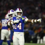 FootballR - NFL - Ein Footballspieler, Stefon Diggs, im Trikot mit der Nummer 84 gestikuliert während eines Nachtspiels auf dem Feld, während im Hintergrund seine Teamkollegen unter hellen Stadionlichtern nach einem Blockbuster-Trade zu sehen sind. Diese Beschreibung wurde automatisch generiert.