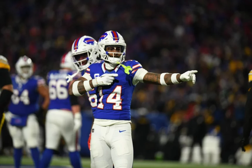 FootballR - NFL - Ein Footballspieler, Stefon Diggs, im Trikot mit der Nummer 84 gestikuliert während eines Nachtspiels auf dem Feld, während im Hintergrund seine Teamkollegen unter hellen Stadionlichtern nach einem Blockbuster-Trade zu sehen sind. Diese Beschreibung wurde automatisch generiert.