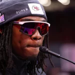 FootballR - NFL - Eine Person, Rashee Rice, die eine Mütze mit dem Super Bowl-Logo und eine Sonnenbrille trägt, spricht in ein Mikrofon, zieht eine Chiefs WR-Jacke an und deutet damit auf einen Athleten bei einer Presseveranstaltung hin. Diese Beschreibung wurde automatisch generiert.