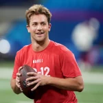 FootballR - NFL - Ein junger Mann mit zerzaustem Haar lächelt breit und hält einen Football in einer Sporthalle. Er trägt ein rotes T-Shirt mit dem Aufdruck „Novill Free Agent“ und der Nummer 12 in Weiß. Diese Beschreibung wurde automatisch generiert. Kedon Slovis