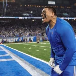 FootballR - NFL - Penei Sewell - Ein Footballspieler, wahrscheinlich ein Offensive Tackle der Lions, in einer blauen Uniform feiert leidenschaftlich am Spielfeldrand eines voll besetzten Stadions, in der Nähe der hellblauen Endzone, während die Menge im Hintergrund erscheint Diese Beschreibung wurde automatisch generiert.