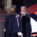 FootballR - NFL - Zwei Männer lächeln auf einer Bühne mit einem großen, orange-blauen Sport-Hintergrund. Caleb Williams in einem marineblauen Anzug und einer Mütze schüttelt einem älteren Mann im Anzug die Hand und feiert einen bedeutsamen Diese Beschreibung wurde automatisch generiert.