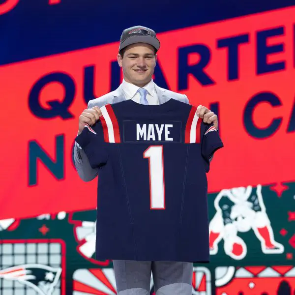 FootballR - NFL - Ein junger Mann auf einer Bühne beim NFL Draft 2024, lächelt und hält ein Footballtrikot mit Drake Maye und der Nummer „1“ hoch. Ein leuchtend roter und blauer Hintergrund Diese Beschreibung wurde automatisch generiert.